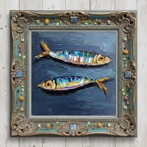 May include: An oil painting of two fish with iridescent blue, yellow, and silver scales swimming against a dark navy blue background. The artwork is encased in an ornate, weathered teal and gold frame decorated with small floral and tile accents.