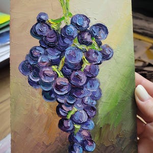 Oil painting of a bunch of grapes in shades of purple and blue, with green stems and leaves. The artwork is on a 5x7 inch panel, with a textured brushstroke style. The background has a mix of brown and green tones.
