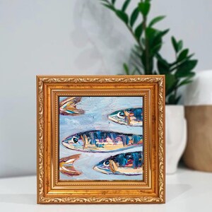 May include: Square framed oil painting featuring a school of fish. The fish are painted with blue, orange, and yellow brushstrokes against a light blue background. The frame is gold with ornate detailing.