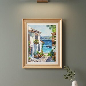 May include: A framed oil painting depicting a coastal scene with a white Mediterranean-style building, potted plants, and a stone path leading to a bright blue sea under a clear sky. The artwork is enclosed in a gold-toned frame with a white mat.
