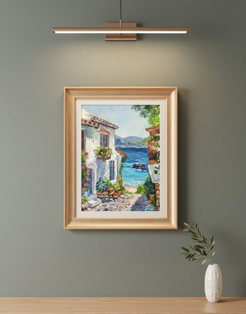 May include: A framed oil painting depicting a coastal scene with a white Mediterranean-style building, potted plants, and a stone path leading to a bright blue sea under a clear sky. The artwork is enclosed in a gold-toned frame with a white mat.