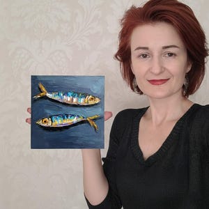 May include: A square oil painting on canvas featuring two fish with iridescent scales in shades of blue, yellow, and silver, set against a dark, textured navy blue background with visible brushstrokes throughout the entire composition.