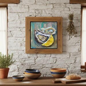 May include: A framed painting of oysters and a lemon slice. The artwork is in a wooden frame and hangs on a white brick wall. A small potted plant, bowls, and a loaf of bread are on a wooden surface.