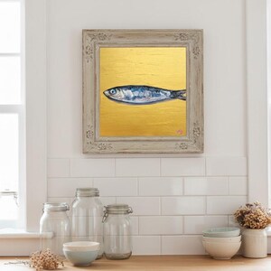 May include: Framed artwork featuring a blue and silver fish painted on a gold background. The painting is in a distressed, light-colored wooden frame. The artwork is displayed above a wooden countertop with jars, bowls, and a vase.