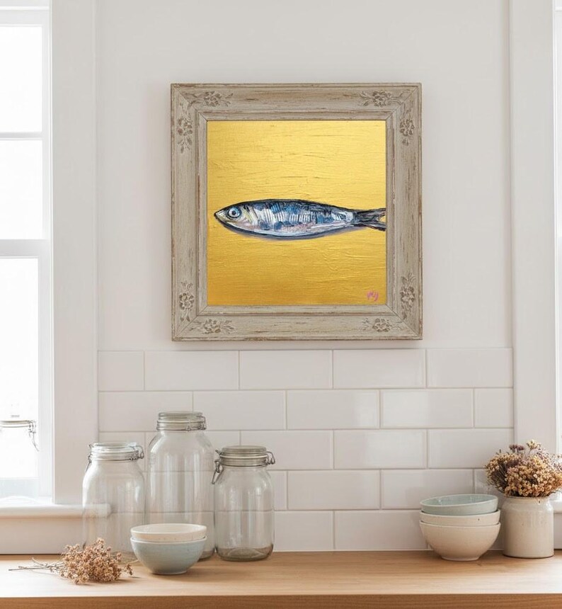 May include: Framed artwork featuring a blue and silver fish painted on a gold background. The painting is in a distressed, light-colored wooden frame. The artwork is displayed above a wooden countertop with jars, bowls, and a vase.