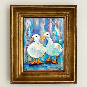 May include: Oil painting of two white ducks with orange beaks and feet, set against a colorful background of blues, purples, and turquoise. The artwork is framed in a gold-colored, ornate frame, adding a touch of elegance to the piece. The painting is signed in the bottom right corner.
