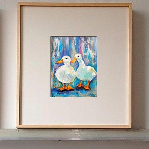 May include: An oil painting of two white ducks with orange beaks and feet, set against a blue and purple background. The artwork is framed in a light wood frame and matted with a white border.