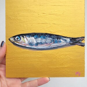 A small painting of a silver fish on a gold background. The fish has a blue eye and is painted in a realistic style.