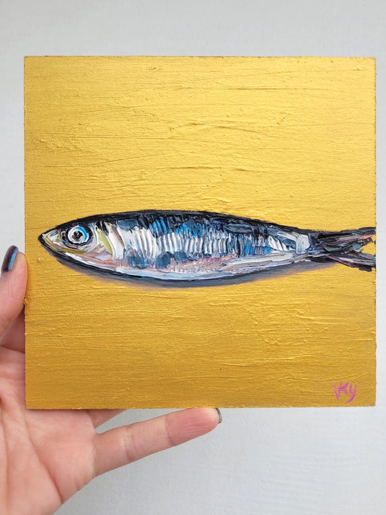 A small painting of a silver fish on a gold background. The fish has a blue eye and is painted in a realistic style.
