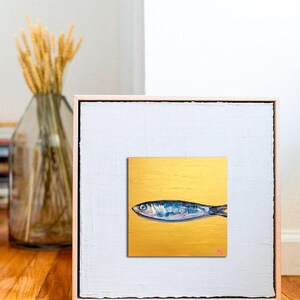 May include: A framed artwork featuring a stylized painting of a fish. The fish is depicted in shades of blue and silver against a gold background. The artwork is encased in a white frame with a light wood border, creating a modern aesthetic.