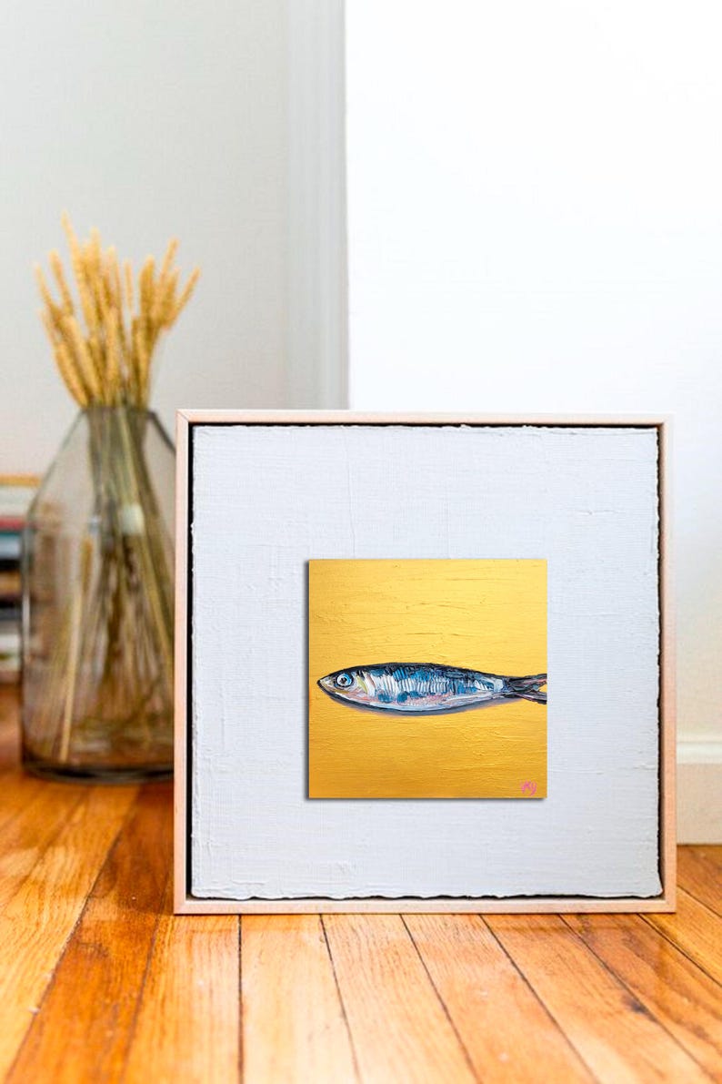 May include: A framed artwork featuring a stylized painting of a fish. The fish is depicted in shades of blue and silver against a gold background. The artwork is encased in a white frame with a light wood border, creating a modern aesthetic.