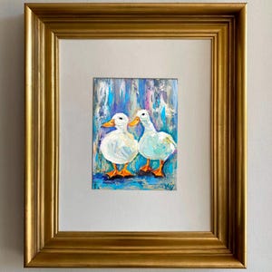 Oil painting of two white ducks with orange feet and beaks, set against a blue and white background. The artwork is framed in a gold-colored, ornate frame, creating a decorative piece.