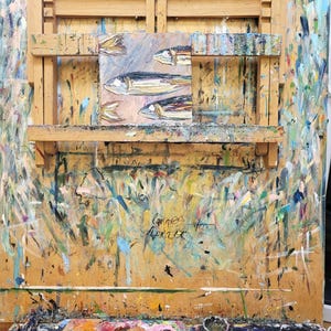 A wooden artist's easel with a small painting of fish on it. The easel is covered in paint splatters and has a paint palette on the bottom.