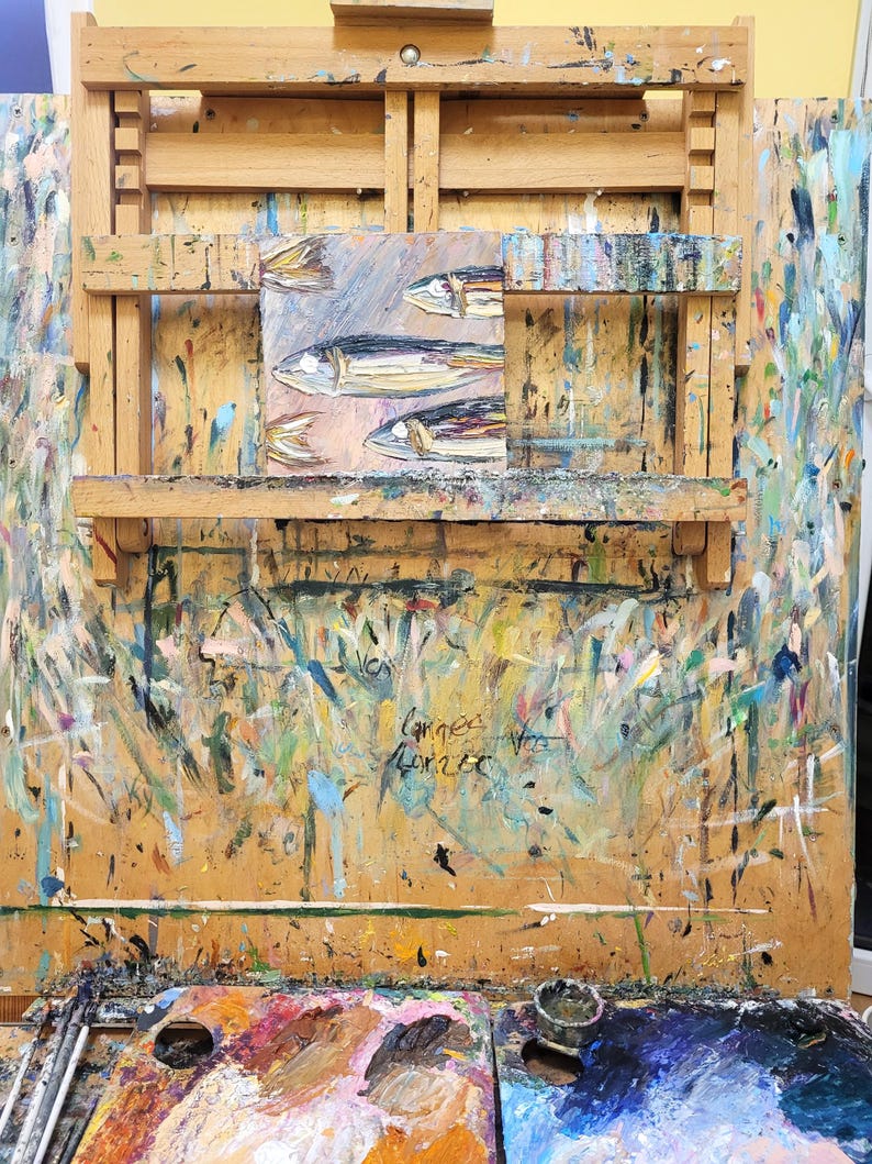 A wooden artist's easel with a small painting of fish on it. The easel is covered in paint splatters and has a paint palette on the bottom.