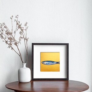 May include: Framed art print featuring a blue and silver fish on a gold background. The artwork is displayed in a black frame with a white mat. The print is on a round, brown table with a white vase and decorative branches.