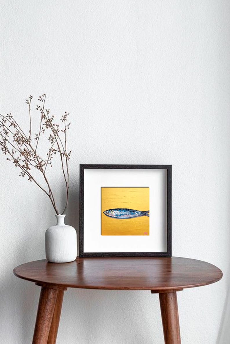 May include: Framed art print featuring a blue and silver fish on a gold background. The artwork is displayed in a black frame with a white mat. The print is on a round, brown table with a white vase and decorative branches.