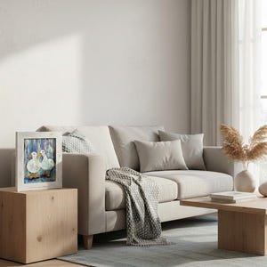 May include: A framed artwork featuring two white ducks with blue and orange accents. The painting is set against a blue background and framed in a distressed white wood. The artwork is displayed on a light wood side table next to a beige sofa.