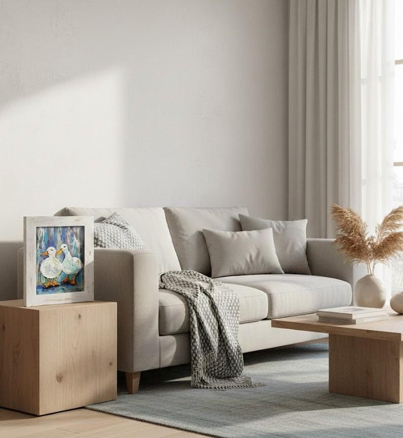 May include: A framed artwork featuring two white ducks with blue and orange accents. The painting is set against a blue background and framed in a distressed white wood. The artwork is displayed on a light wood side table next to a beige sofa.