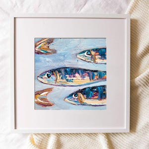 May include: An original oil painting of several fish swimming in a light blue sea. The fish are painted with textured brushstrokes in shades of blue, yellow, and orange. The artwork is framed in a white square frame.