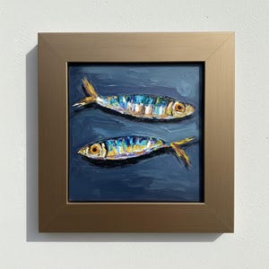 An oil painting of two fish in a dark blue sea. The fish are painted with bright colors, including yellow, blue, and white. The painting is framed in a gold-colored frame.