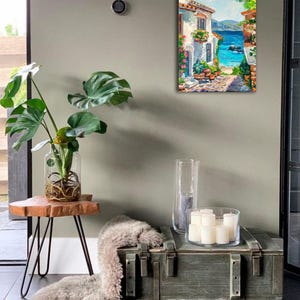 May include: A rectangular oil painting on canvas depicting a Mediterranean coastal scene with a white stone building, vibrant potted flowers, a cobblestone path, and a bright blue sea under a clear sky, displayed in a vertical orientation.