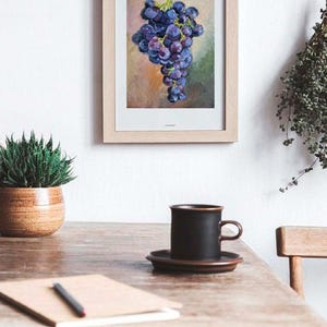 May include: A framed oil painting of a cluster of dark purple grapes hanging from a green stem. The artwork features a textured background with blended shades of tan, pink, and green. The frame is made of light-colored wood with a white mat border.