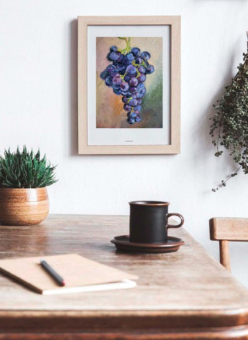 May include: A framed oil painting of a cluster of dark purple grapes hanging from a green stem. The artwork features a textured background with blended shades of tan, pink, and green. The frame is made of light-colored wood with a white mat border.