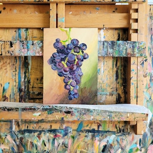 An oil painting of a bunch of grapes, rendered in shades of purple, blue, and green. The artwork is displayed on a wooden easel, with a colorful, paint-splattered surface below. The painting's background features warm tones.