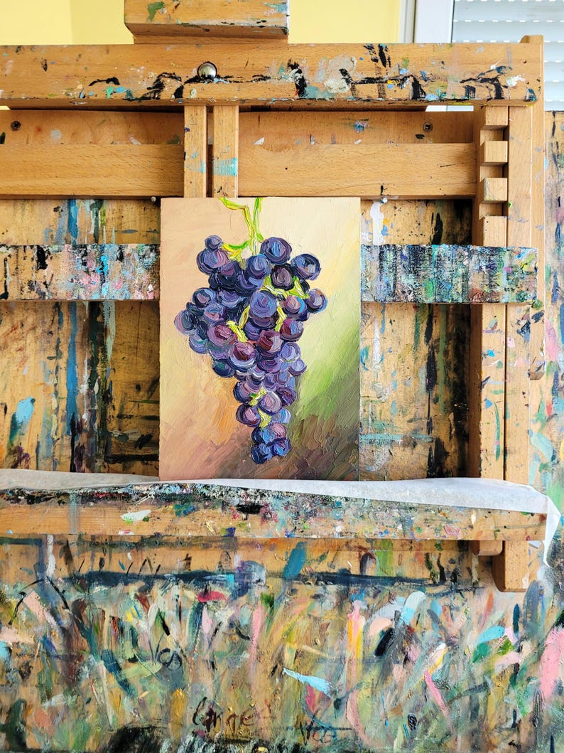 An oil painting of a bunch of grapes, rendered in shades of purple, blue, and green. The artwork is displayed on a wooden easel, with a colorful, paint-splattered surface below. The painting's background features warm tones.