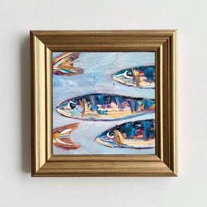 May include: Square framed oil painting of several fish swimming in a light blue sea. The fish are painted with blue, orange, and yellow brushstrokes. The frame is gold.