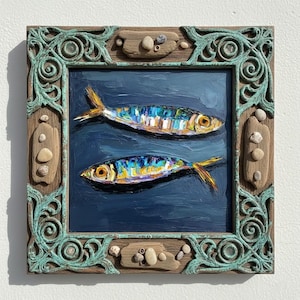 A square, framed painting of two fish. The fish are painted with bright colors, including yellow, blue, and white, against a dark blue background. The frame is ornate, with a teal finish and decorative elements.