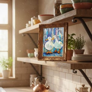 May include: A framed painting of two white ducks with orange feet and beaks, set against a blue and white background. The painting has a gold ornate frame and is displayed on a wooden shelf in a kitchen setting. Other kitchen items are visible.