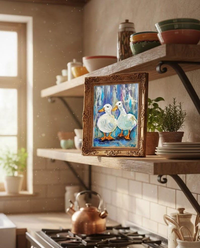 May include: A framed painting of two white ducks with orange feet and beaks, set against a blue and white background. The painting has a gold ornate frame and is displayed on a wooden shelf in a kitchen setting. Other kitchen items are visible.