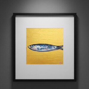 May include: Framed artwork featuring a detailed painting of a fish. The fish is depicted in shades of blue and silver against a textured gold background. The artwork is encased in a black frame with a white mat.
