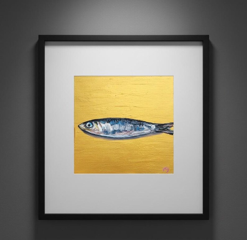 May include: Framed artwork featuring a detailed painting of a fish. The fish is depicted in shades of blue and silver against a textured gold background. The artwork is encased in a black frame with a white mat.