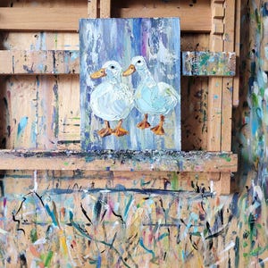 An oil painting on canvas depicts two white ducks with orange beaks and feet. The ducks are set against a background of vertical blue, purple, and white brushstrokes. The artwork is displayed on a wooden easel.