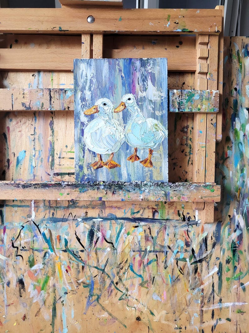 An oil painting on canvas depicts two white ducks with orange beaks and feet. The ducks are set against a background of vertical blue, purple, and white brushstrokes. The artwork is displayed on a wooden easel.