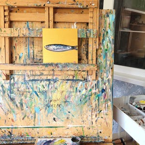 A small painting of a silver fish on a yellow background, displayed on an easel with a paint-splattered surface. The easel is made of wood and has a drawer on the side.