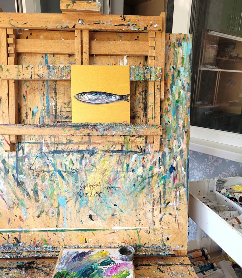 A small painting of a silver fish on a yellow background, displayed on an easel with a paint-splattered surface. The easel is made of wood and has a drawer on the side.