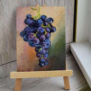May include: An oil painting on a rectangular canvas depicting a cluster of dark purple and blue grapes with green stems. The artwork is displayed on a small, light-colored wooden easel. The background of the painting features blended shades of pink, orange, and dark green.