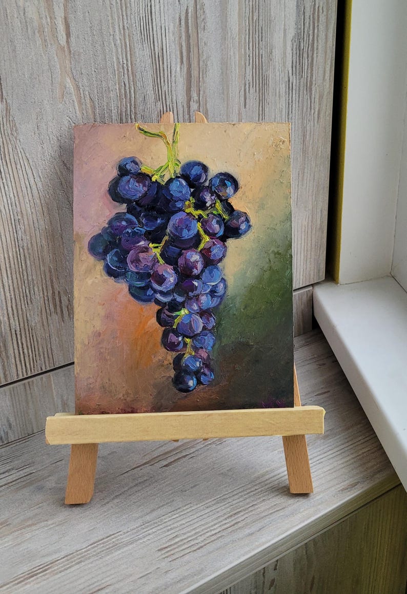 May include: An oil painting on a rectangular canvas depicting a cluster of dark purple and blue grapes with green stems. The artwork is displayed on a small, light-colored wooden easel. The background of the painting features blended shades of pink, orange, and dark green.