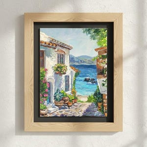 May include: A framed oil painting depicting a Mediterranean coastal scene with a white stucco building, vibrant potted flowers, a stone pathway, and a view of the blue sea under a bright sky, all enclosed in a light-toned wooden frame with a black mat.