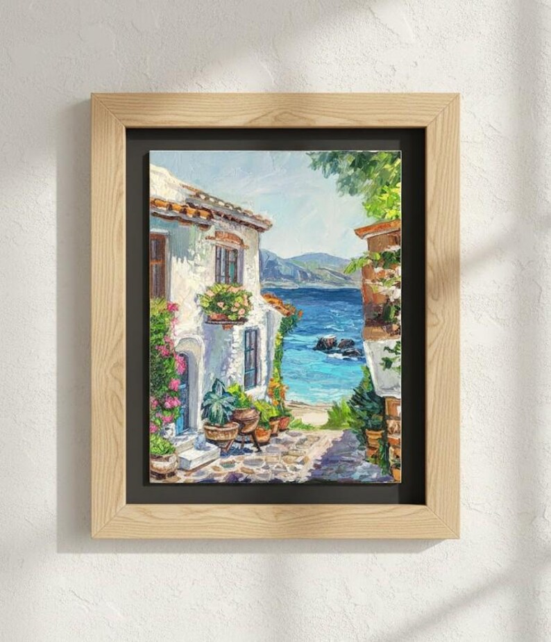 May include: A framed oil painting depicting a Mediterranean coastal scene with a white stucco building, vibrant potted flowers, a stone pathway, and a view of the blue sea under a bright sky, all enclosed in a light-toned wooden frame with a black mat.