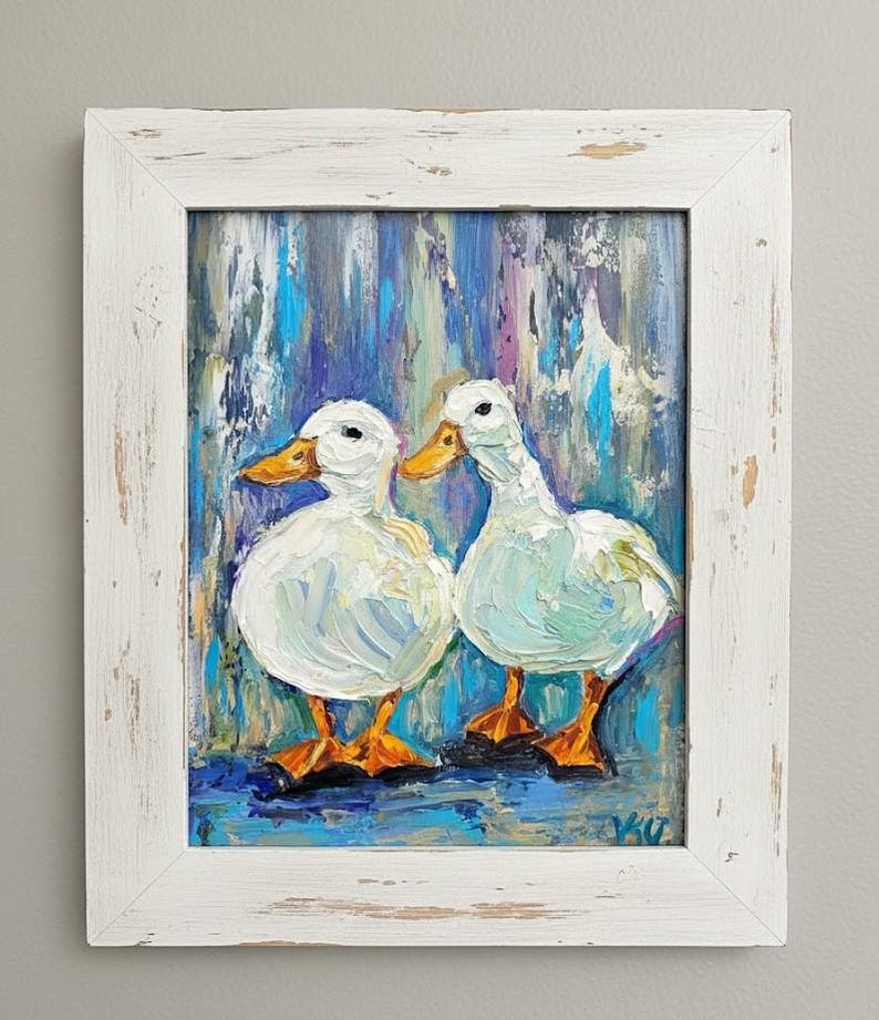 May include: Oil painting of two white ducks with orange beaks and feet, set against a blue, purple, and teal background. The artwork is framed in a distressed white wooden frame, adding a rustic touch to the piece.