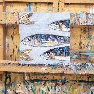 May include: An original oil painting of several fish, rendered in a textured style with shades of blue, brown, and white. The artwork is displayed on a wooden easel, showcasing the artist's work.