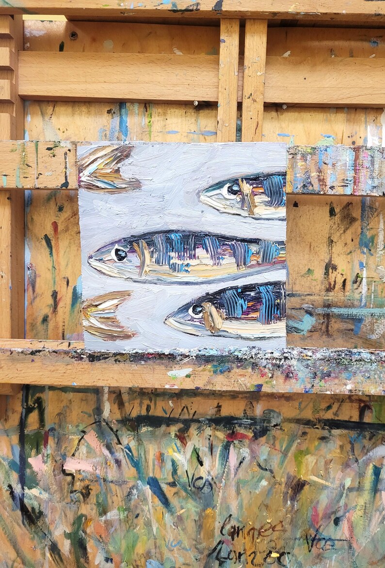 May include: An original oil painting of several fish, rendered in a textured style with shades of blue, brown, and white. The artwork is displayed on a wooden easel, showcasing the artist's work.