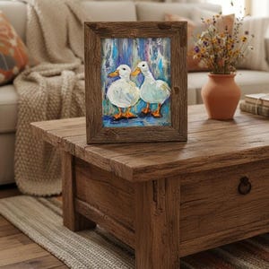 May include: A framed painting of two white ducks with orange feet and beaks. The artwork is set against a blue and white background. The frame is made of rustic brown wood. The painting is displayed on a wooden coffee table.