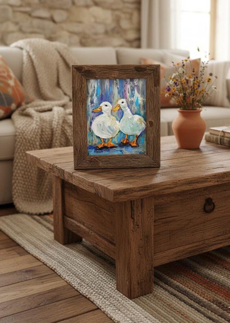 May include: A framed painting of two white ducks with orange feet and beaks. The artwork is set against a blue and white background. The frame is made of rustic brown wood. The painting is displayed on a wooden coffee table.