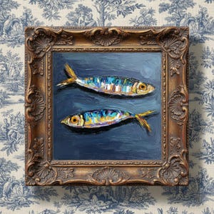 May include: An oil painting featuring two fish with iridescent blue, yellow, and silver scales against a dark navy background. The artwork is encased in an ornate, antique-style gold frame with intricate carved detailing along the edges.