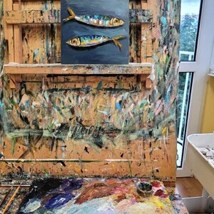 Oil painting of two fish with colorful scales and yellow fins on a dark blue background. The artwork is displayed on an artist's easel, surrounded by paint splatters and a colorful palette, suggesting a studio setting.